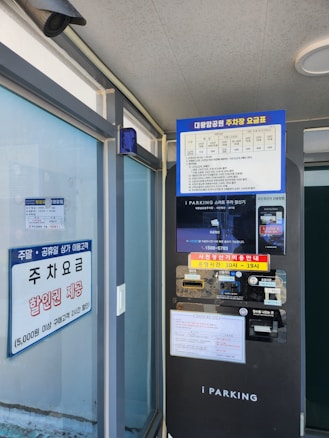 A parking payment kiosk with a screen displaying information in Korean. The kiosk includes a payment terminal and various instructions. Adjacent to the kiosk is a glass window with signage about parking fees and discounts. An overhead security camera is visible.