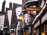 A quaint, old-fashioned street with narrow, tall buildings featuring snow-dusted roofs and glass windows displaying various items. Warm, glowing lanterns light up the street, adding a cozy atmosphere. The sign 'SCRIVENSHAFT'S Finest Parchments, Inks & Quills' is clearly visible, indicating a shop specializing in stationery supplies.