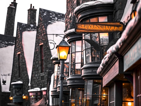 A quaint, old-fashioned street with narrow, tall buildings featuring snow-dusted roofs and glass windows displaying various items. Warm, glowing lanterns light up the street, adding a cozy atmosphere. The sign 'SCRIVENSHAFT'S Finest Parchments, Inks & Quills' is clearly visible, indicating a shop specializing in stationery supplies.