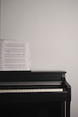 A black piano with a sheet of music placed on its open cover against a plain white wall.