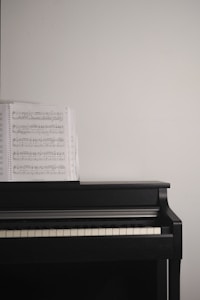 A black piano with a sheet of music placed on its open cover against a plain white wall.
