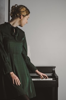 A woman in a dark green dress is leaning towards a piano, appearing to be lightly touching the keys. Her hair is styled in an elegant updo, and she is gazing downwards.