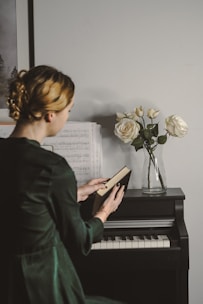 A person with blonde hair is seated at a piano, holding an open book. Sheet music is placed on the piano stand, and a glass vase with white roses is positioned on top of the piano. The atmosphere is calm and introspective.