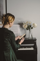 A person with blonde hair is seated at a piano, holding an open book. Sheet music is placed on the piano stand, and a glass vase with white roses is positioned on top of the piano. The atmosphere is calm and introspective.