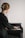 A person with blonde hair is playing a piano while reading sheet music. They are wearing a dark green garment, and their hair is styled with a large gold clip. The piano is a small upright model, and the lighting is soft, creating a serene and focused atmosphere.