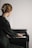 A person with blonde hair is playing a piano while reading sheet music. They are wearing a dark green garment, and their hair is styled with a large gold clip. The piano is a small upright model, and the lighting is soft, creating a serene and focused atmosphere.