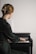 A person with blonde hair is playing a piano while reading sheet music. They are wearing a dark green garment, and their hair is styled with a large gold clip. The piano is a small upright model, and the lighting is soft, creating a serene and focused atmosphere.
