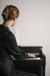 A person with blonde hair is playing a piano while reading sheet music. They are wearing a dark green garment, and their hair is styled with a large gold clip. The piano is a small upright model, and the lighting is soft, creating a serene and focused atmosphere.