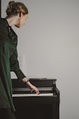 A person dressed in a dark green outfit stands beside a black piano, gently resting their hand on the keys. Sheet music is placed on the piano, and the person's head is turned to the side, suggesting focus or contemplation.