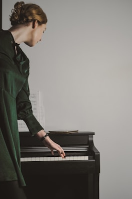 A person dressed in a dark green outfit stands beside a black piano, gently resting their hand on the keys. Sheet music is placed on the piano, and the person's head is turned to the side, suggesting focus or contemplation.
