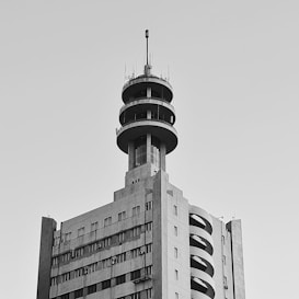 A tall, rectangular building with multiple floors and a distinctive circular tower on top, featuring various antennas. The facade includes vertical and horizontal lines indicative of windows and other architectural features.