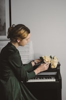 A person with styled hair and wearing a dark green dress is sitting at a piano. Sheet music is propped up on the piano, and the person is holding a bouquet of pale yellow roses. The scene is softly lit, creating a calm atmosphere.