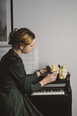 A person with styled hair and wearing a dark green dress is sitting at a piano. Sheet music is propped up on the piano, and the person is holding a bouquet of pale yellow roses. The scene is softly lit, creating a calm atmosphere.