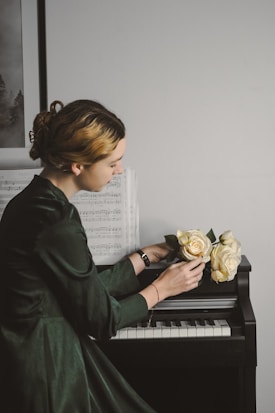 A person with styled hair and wearing a dark green dress is sitting at a piano. Sheet music is propped up on the piano, and the person is holding a bouquet of pale yellow roses. The scene is softly lit, creating a calm atmosphere.