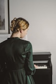 A person with pinned back blonde hair wearing an elegant dark green dress stands in front of a piano. They are positioned with their back to the viewer, gently touching the piano keys. Sheet music is visible in the background, resting against the wall.