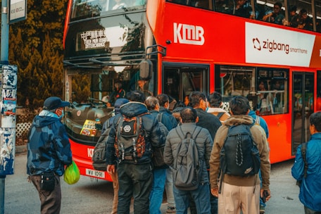 A group of people queue at a bus stop, waiting to board a red double-decker bus. The bus has signage for 'Tuen Mun Road Interchange' and various advertisements. Many individuals wear jackets and carry backpacks, indicating a casual and everyday setting. Some people are wearing face masks.