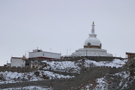 A large white stupa with intricate designs is situated on a snow-covered hillside. Surrounding the stupa are smaller structures, with a mix of modern and traditional architecture. The sky is overcast, contributing to the muted color palette of the landscape.