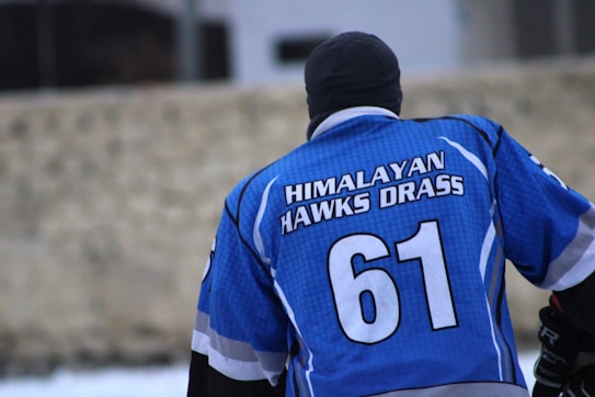 A person is seen from behind wearing a blue sports jersey with the text 'Himalayan Hawks Drass' and the number 61. The person is also wearing a black beanie. The background appears to be an outdoor setting.