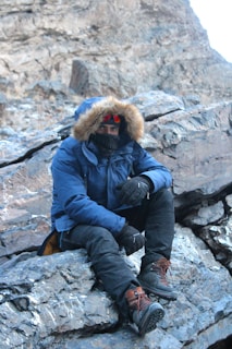 A person wearing a fur-lined winter jacket, thick boots, and gloves is seated on a rocky surface in a mountainous or rugged terrain. The individual is also equipped with a face covering or scarf and tinted goggles, indicating cold weather conditions.