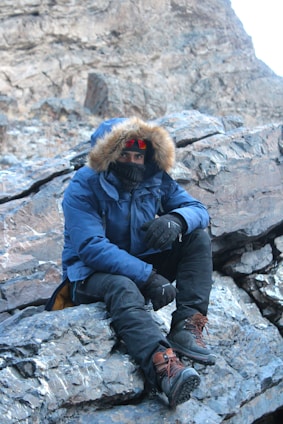 A person wearing a fur-lined winter jacket, thick boots, and gloves is seated on a rocky surface in a mountainous or rugged terrain. The individual is also equipped with a face covering or scarf and tinted goggles, indicating cold weather conditions.