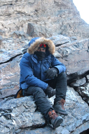 A person wearing a fur-lined winter jacket, thick boots, and gloves is seated on a rocky surface in a mountainous or rugged terrain. The individual is also equipped with a face covering or scarf and tinted goggles, indicating cold weather conditions.