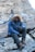 A person wearing a fur-lined winter jacket, thick boots, and gloves is seated on a rocky surface in a mountainous or rugged terrain. The individual is also equipped with a face covering or scarf and tinted goggles, indicating cold weather conditions.