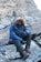 A person wearing a fur-lined winter jacket, thick boots, and gloves is seated on a rocky surface in a mountainous or rugged terrain. The individual is also equipped with a face covering or scarf and tinted goggles, indicating cold weather conditions.