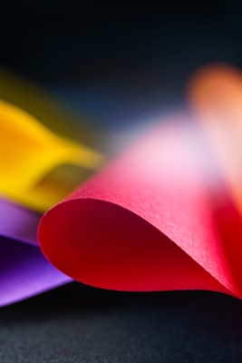 Curved sheets of colorful paper create an abstract and vibrant composition against a dark background. The focus is on a red sheet in the foreground with hints of purple and yellow visible in the background.