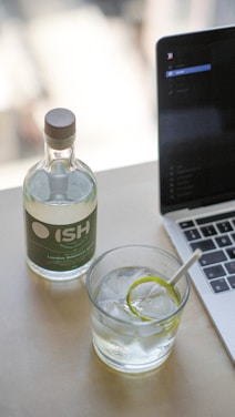 A bottle with a green label labeled 'ISH' stands next to a glass filled with a clear liquid, ice cubes, and a slice of lime on a table. Beside it, a laptop is partially visible, indicating a workspace setting.