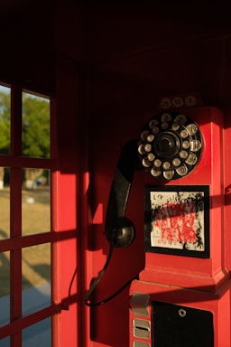 An old-fashioned rotary payphone with a black handset is mounted inside a red telephone booth. The booth's interior is illuminated by warm sunlight, casting shadows on the red walls. Outside, a glimpse of grass and a tree is visible.