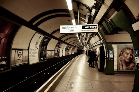 A London Underground station platform with curved walls adorned with various advertisements. Signs indicate directions for exits and lifts. A few people are seen waiting near the edge of the platform. The lighting is subdued, creating a vintage look.