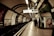 A London Underground station platform with curved walls adorned with various advertisements. Signs indicate directions for exits and lifts. A few people are seen waiting near the edge of the platform. The lighting is subdued, creating a vintage look.
