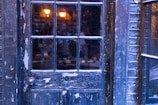 A weathered, dark blue wooden door with peeling paint and windowpanes reflects faint interior lighting. Warm glowing lights are visible through the window, casting a cozy ambiance, while exterior brickwork surrounds the door frame.