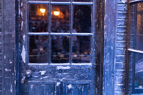 A weathered, dark blue wooden door with peeling paint and windowpanes reflects faint interior lighting. Warm glowing lights are visible through the window, casting a cozy ambiance, while exterior brickwork surrounds the door frame.