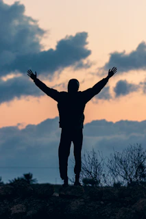 A silhouette of a person standing strong on a rocky path, arms raised in worship against a glowing horizon.