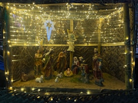 A nativity scene featuring figurines of the Holy Family along with the Magi and shepherds inside a stable. The scene is decorated with warm yellow string lights and a large star hangs above.