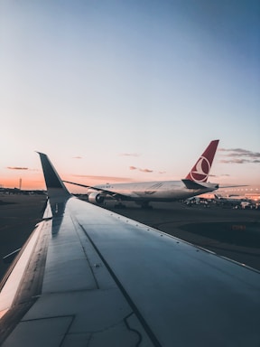 An airport scene at sunset featuring the wing of a passenger plane in the foreground, with another airplane in the background displaying a distinct red tail fin with a circular logo. The sky is tinged with warm hues as the sun sets, and airport lights and infrastructure can be seen faintly in the distance.