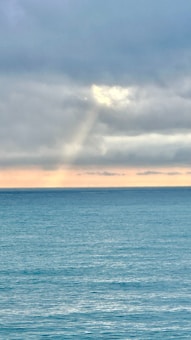 A serene seascape with calm waters under a cloud-covered sky. The horizon displays a subtle orange glow, suggesting a sunrise or sunset. A beam of light breaks through the clouds, casting a gentle reflection on the water.
