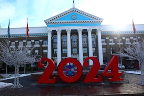 In front of a neoclassical building with white columns and a blue pediment, large red numbers reading '2024' are prominently displayed. There are two bare trees adorned with white lights and flags on poles nearby. The sun is shining brightly from the right corner, creating a dynamic lighting effect.