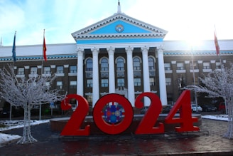 In front of a neoclassical building with white columns and a blue pediment, large red numbers reading '2024' are prominently displayed. There are two bare trees adorned with white lights and flags on poles nearby. The sun is shining brightly from the right corner, creating a dynamic lighting effect.