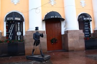 An outdoor scene of a building with yellow walls and large white columns, featuring two black signs with cyrillic writing and a list of words in English such as 'smile', 'eat', 'laugh', 'talk', 'dance', and 'drink'. A metal sculpture of a man wearing a hat and suit stands in front, with a wreath decoration on the wooden door behind it. The area includes festive decorations and references to live music.