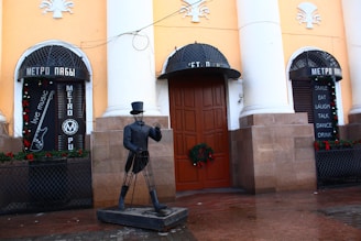 An outdoor scene of a building with yellow walls and large white columns, featuring two black signs with cyrillic writing and a list of words in English such as 'smile', 'eat', 'laugh', 'talk', 'dance', and 'drink'. A metal sculpture of a man wearing a hat and suit stands in front, with a wreath decoration on the wooden door behind it. The area includes festive decorations and references to live music.