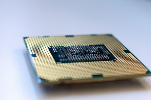 A close-up view of a computer processor chip lying on a surface, showing its intricate gold-colored pins and patterns. The focus is on the detailed grid-like design, with slight blurring at the edges.