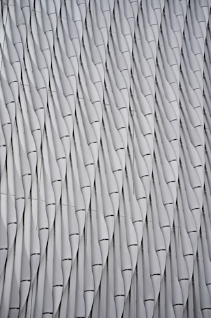 A repeating pattern of abstract, wavy metallic panels creating an architectural texture. The panels are intricately designed and aligned in vertical rows, forming a series of gentle curves and shadows.