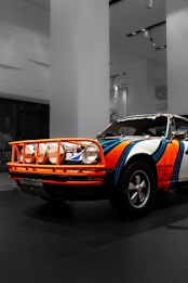 A vintage rally car with a colorful paint job is parked indoors. It features bold stripes in blue and orange, with prominent sponsorship decals on the side. The car is fitted with additional spotlights on the front and has classic tires.