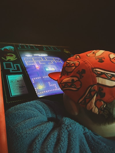 A person wears a vibrant red cap decorated with cartoon-style graphics, leaning over a classic arcade game screen. The retro game interface is brightly lit, featuring iconic maze and dot patterns, with Pac-Man graphics and glowing colors.