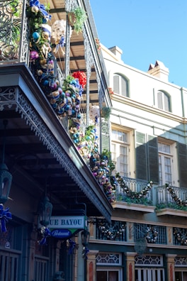 The image features a charming urban scene with intricately designed wrought iron balconies decorated with colorful ornaments and garlands. The buildings have a classic architectural style with pastel colors and shuttered windows. A sign for a restaurant named 'Blue Bayou' is visible, adding to the festive ambiance.