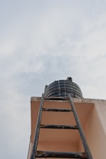 Looking up towards a black water tank sitting atop a building alongside a dark metal ladder leading up to it. The scene is set against a backdrop of a cloudy sky.