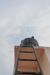 Looking up towards a black water tank sitting atop a building alongside a dark metal ladder leading up to it. The scene is set against a backdrop of a cloudy sky.