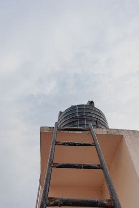 Looking up towards a black water tank sitting atop a building alongside a dark metal ladder leading up to it. The scene is set against a backdrop of a cloudy sky.